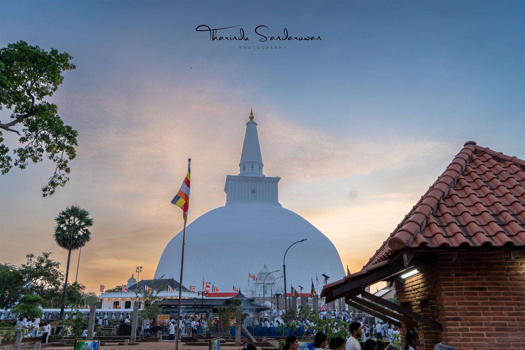 A white stupa at dusk, with a tiled roof and visitors in the foreground