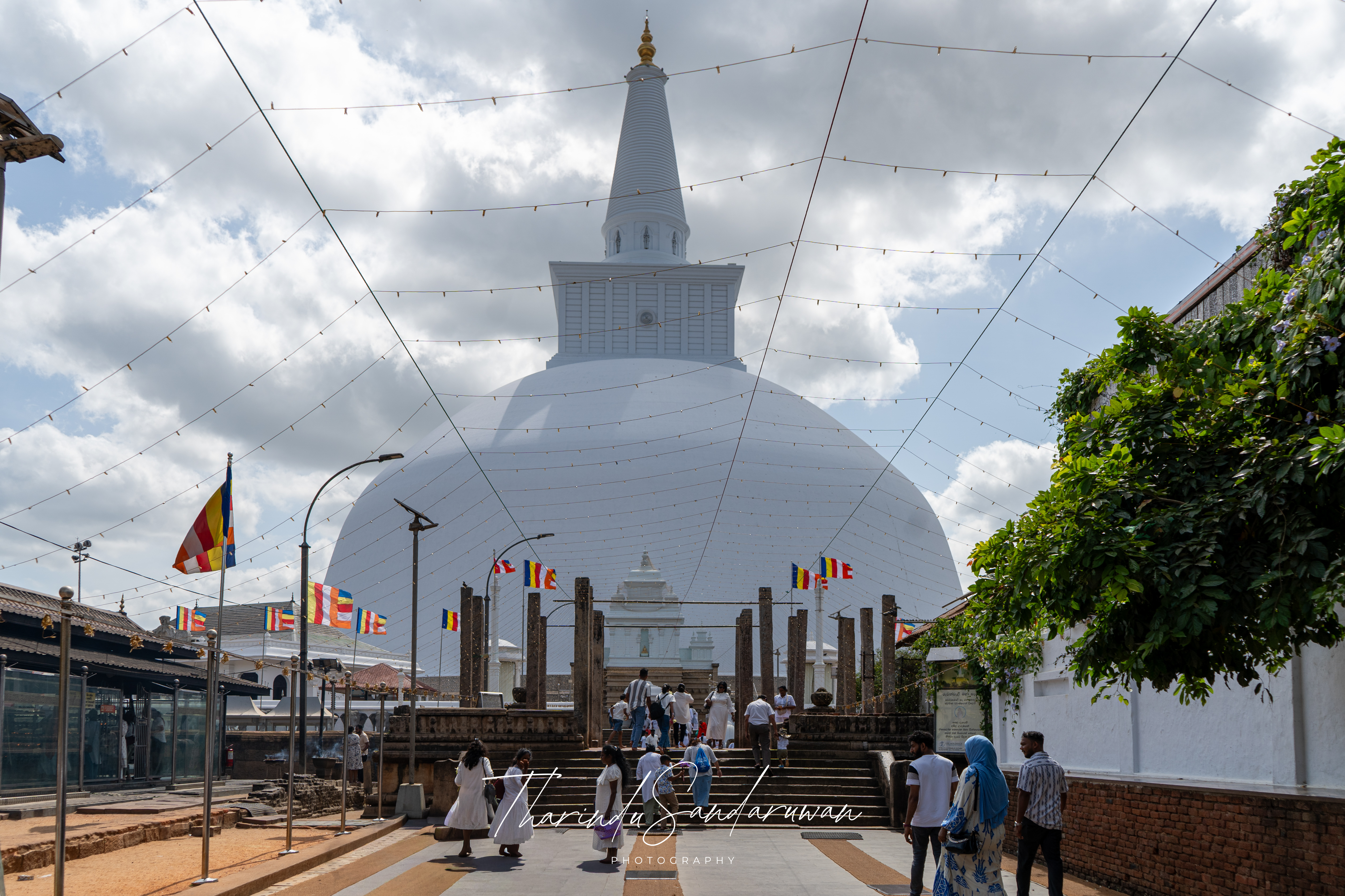 A large white stupa, prayer flags, trees, and visitors under a cloudy sky.