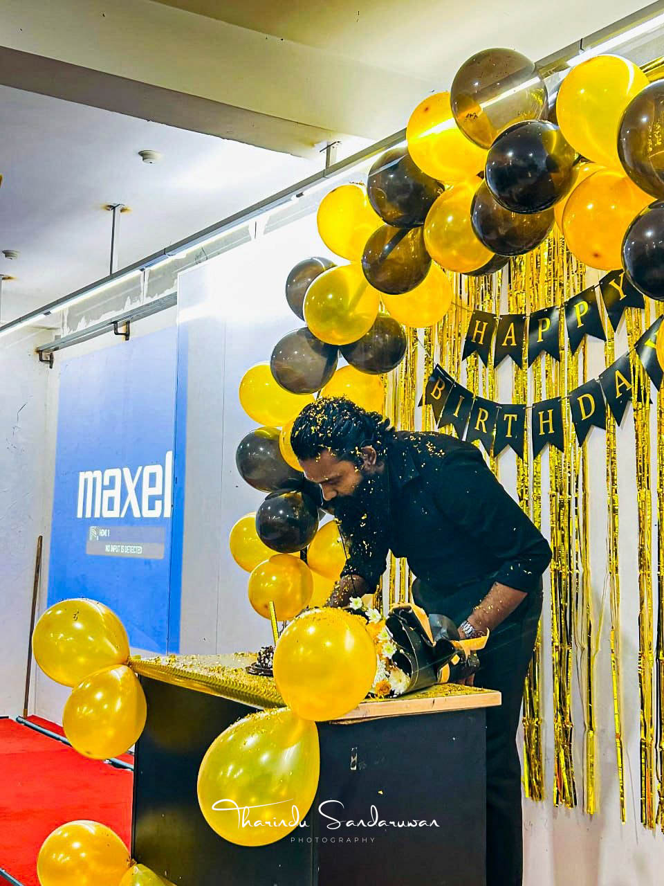 A man cuts a birthday cake surrounded by gold and black balloons.