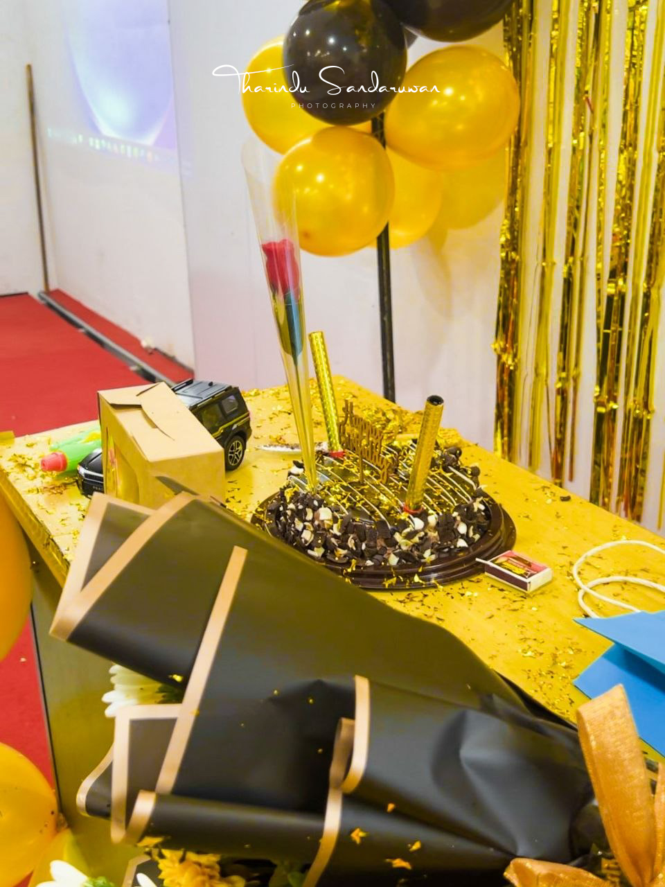 A decorated birthday cake sits on a table beneath gold and black balloons.