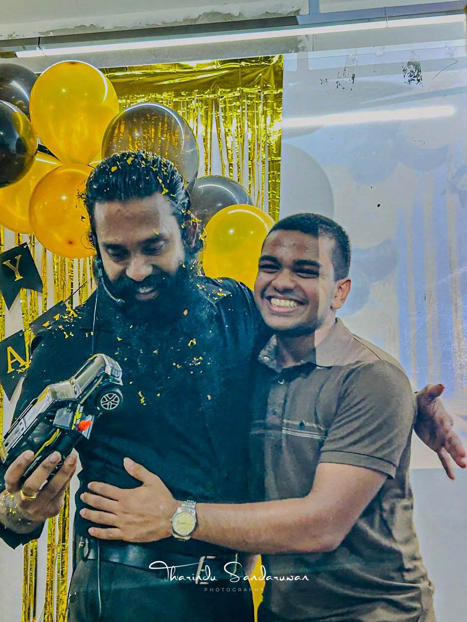 Two men smile together amid balloons and birthday decorations.
