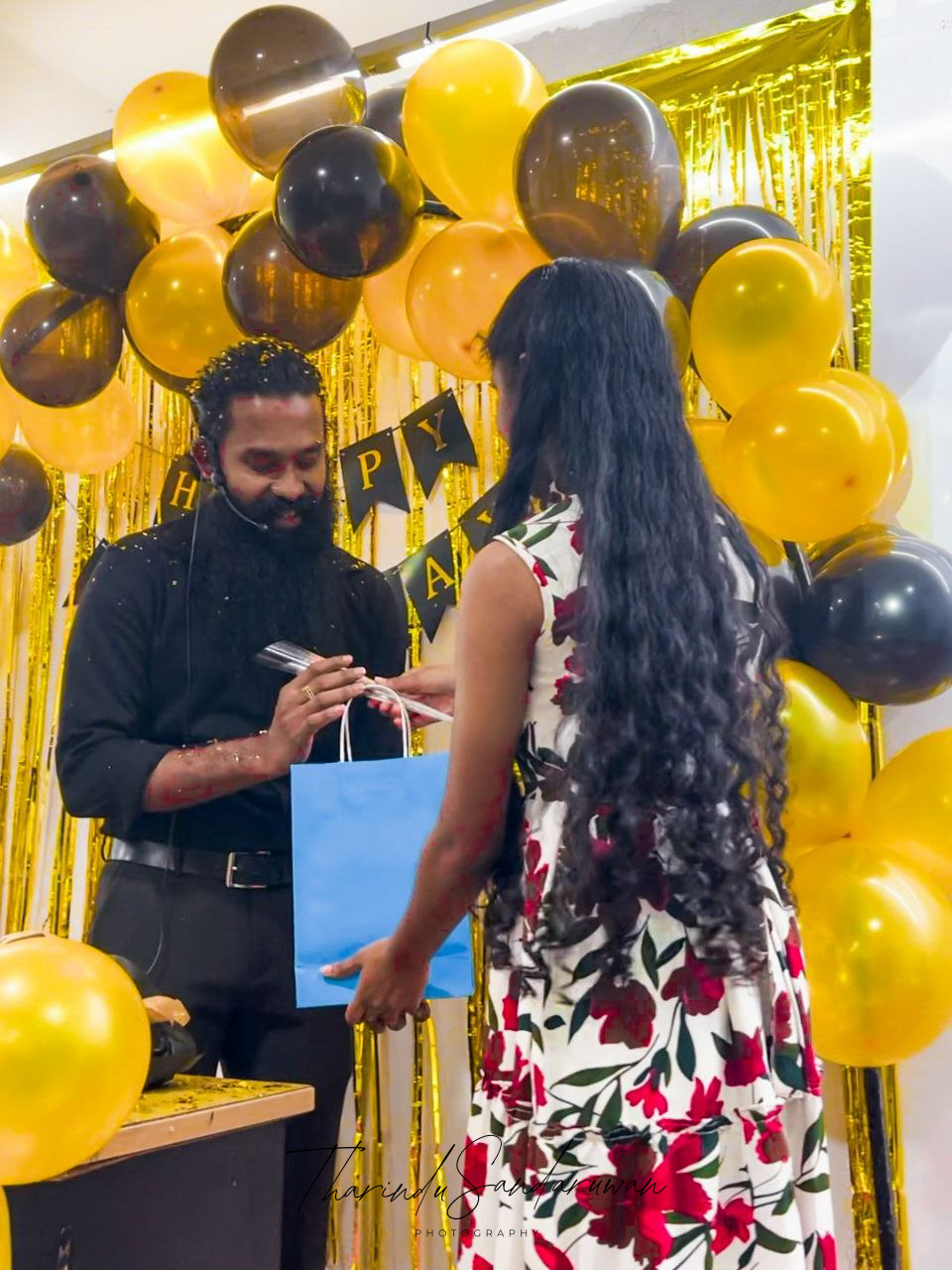 A man and woman exchange a gift bag during a birthday celebration decorated with black and gold balloons.